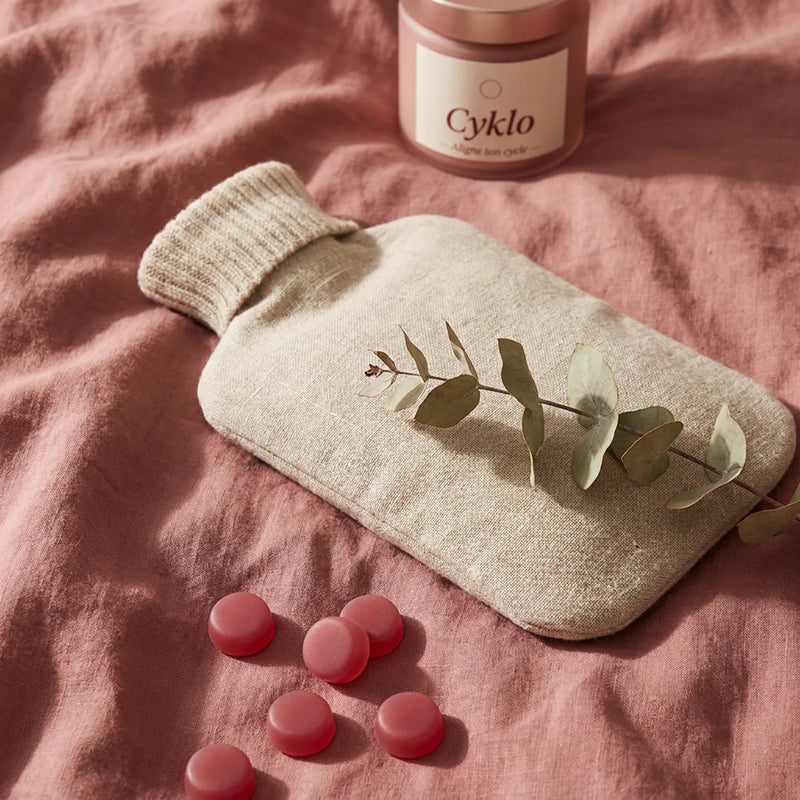 Bouillotte et gummies Cyklo Days sur lit rose nude, solution naturelle contre le mal de ventre pendant les règles